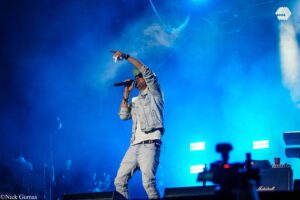 Male singer on stage in a light denim jacket, microphone in hand, pointing to the crowd under blue stage lights.