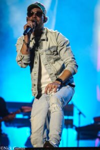 Male singer performing on stage into a microphone, wearing a light denim jacket, ripped jeans, cap, and sunglasses under blue stage lights