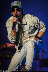Male singer wearing a colorful floral cap and sunglasses, holding a microphone on stage in a light denim jacket and ripped jeans.