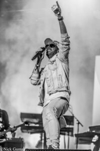 Male singer on stage raising his right fist while singing into a microphone; wearing a denim jacket, cap, and sunglasses (black-and-white photo).