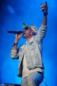 Male singer on stage in a denim jacket and colorful cap, holding a mic and raising a towel- wrapped hand against blue lights.