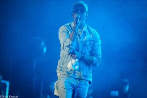 Male singer performing on stage in blue lighting, wearing sunglasses and a denim jacket, singing into a handheld microphone.