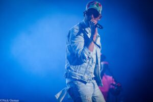 Male singer wearing a floral cap, sunglasses, and denim jacket performs into a microphone on a blue-lit stage.