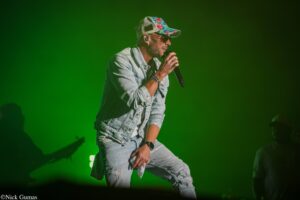 Male singer in a light denim jacket and a colorful floral cap performs on a green-lit stage, gripping a microphone.