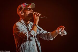 Male singer on a dark stage, mic in hand, wearing a colorful cap and sunglasses under red lighting.