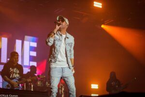 Male singer in a light denim jacket and cap, singing into a handheld mic on a brightly lit stage with orange lights behind him.