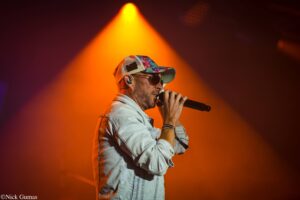 Male singer wearing a floral-pattern cap and denim jacket, singing into a microphone under orange stage lights.
