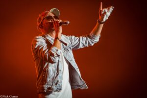 Male singer on stage in denim jacket and cap, sunglasses, microphone in hand, raising a white cloth toward the crowd amid red lighting.