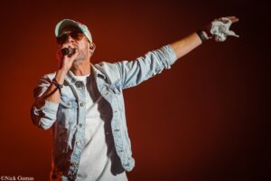 Male singer in a light denim jacket and cap sings into a microphone, one arm extended outward on a red-lit stage.