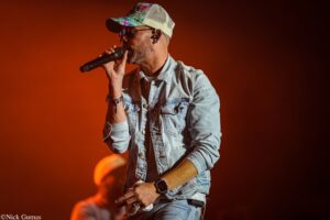 Male singer on an orange-lit stage, wearing a denim jacket and floral cap, singing into a handheld microphone.