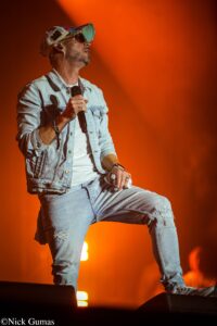 Male singer in a light denim jacket holding a microphone on stage against an orange backdrop, wearing sunglasses and a patterned cap.
