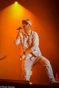 Male singer performing on dark stage with orange spotlight, wearing a denim jacket, cap, sunglasses, and holding a microphone.