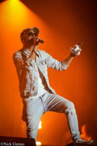 Male singer performing on stage with orange lighting, wearing a denim jacket and sunglasses, holding a microphone.