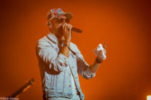 Male singer in a denim jacket and sunglasses performs on an orange-lit stage, holding a microphone.