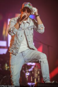 Male singer performing on stage in a denim jacket and tropical cap, holding a microphone and wiping sweat with a cloth.