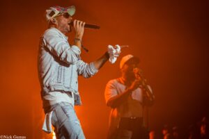Male singer in a denim jacket and colorful cap sings into a microphone on an orange-lit stage, with a towel in hand and band members in the background.