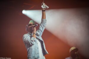 Male performer on stage raises a clenched fist with a white towel, wearing a denim jacket, green cap, and sunglasses under stage lights