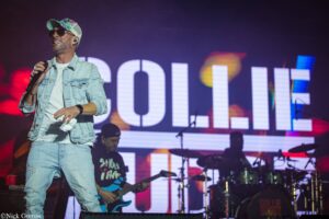 Male singer wearing a light denim jacket and cap, singing into a microphone on a stage with a band in the background