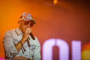 Male vocalist performing on a stage with a microphone, wearing a denim jacket, sunglasses, and a colorful floral cap under warm orange lighting.",
