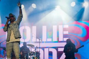 Male singer in a denim jacket and cap performs on a bright concert stage, raising his right fist into the air.