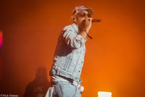 Male singer in a denim jacket and floral cap performing on stage with orange lighting behind him, microphone in hand.