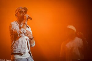 Male singer wearing sunglasses and a denim jacket performs into a microphone on an orange-lit stage.