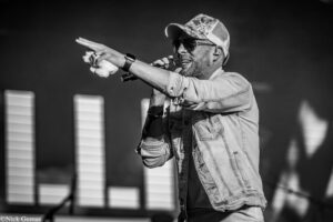 Male singer performing on stage, wearing a cap and sunglasses, holding a microphone and pointing toward the crowd with a towel in hand (black and white).