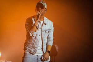 Male singer on stage in a light denim jacket, wearing a patterned cap and sunglasses, singing into a handheld mic with orange lighting behind him.