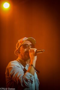Male singer wearing sunglasses and a floral cap, holding a microphone on a dimly lit orange stage.