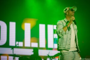 Male singer on stage under vivid green lighting, holding a microphone and wearing a denim jacket and sunglasses.