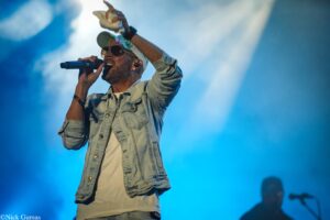Male singer on stage wearing sunglasses and a floral cap, holding a microphone during a live performance under blue lighting