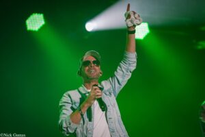 Male performer on stage raising a towel-wrapped hand, wearing a cap and sunglasses, during a concert with green lighting.
