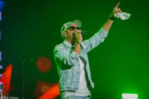 Male vocalist in a denim jacket and cap, wearing sunglasses, singing into a microphone on a green-lit stage with one arm raised and a white cloth in hand.