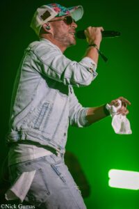 Male singer in a light denim jacket performs on a green-lit stage, holding a microphone and a towel in his other hand, wearing sunglasses and a floral cap.