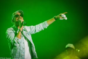 Singer on stage wearing sunglasses and denim jacket, microphone in hand, pointing into the crowd under green lighting.