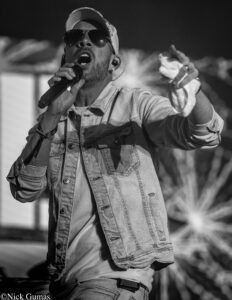 Male singer wearing sunglasses and a cap, holding a microphone while performing on stage.