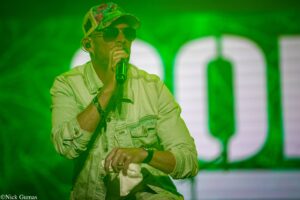 Male singer on stage performing into a microphone, wearing sunglasses and a floral cap under green lighting.