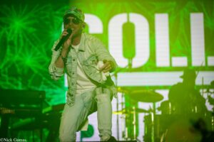 Male singer performing on stage under green lights, holding a microphone and wearing a denim jacket, cap, and sunglasses.