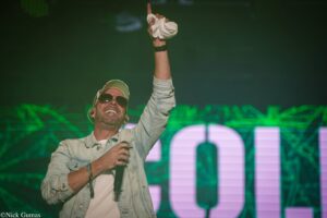Concert performer wearing sunglasses and a denim jacket raises his right arm, clutching a white towel, on a green-lit stage.