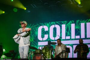 Male singer in a denim jacket and cap performing on a lit stage with a green laser backdrop and band members in the background.