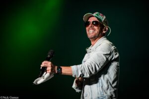 Male singer on stage wearing a denim jacket and sunglasses, holding a microphone with a green spotlight in the background
