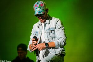 Male singer on stage wearing a colorful tropical cap and denim jacket, smiling while holding a microphone against a green-lit background