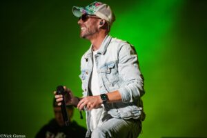 Male singer performing on stage with microphone, wearing a denim jacket, floral cap and sunglasses against a green backdrop.