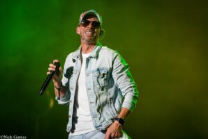 Male singer on stage wearing a denim jacket, white shirt, and sunglasses, holding a microphone and smiling under green lighting.