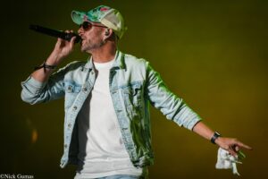 Male singer on stage, mic in hand, wearing a light denim jacket, white tee, and a colorful floral cap with sunglasses photographed mid-performance