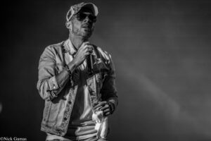 Male singer in a denim jacket and cap, wearing sunglasses, holding a microphone on stage (black and white).