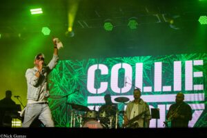 Male singer in a denim jacket and cap performs on a green-lit stage, raising his right hand; band members and a large LED backdrop are visible behind him.