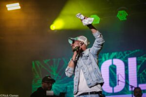 Male singer in a denim jacket and cap raises a white towel while singing into a microphone on a brightly lit stage backgrounded by green and yellow lights.