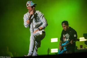 Male singer in a denim jacket and floral cap mid-jump with microphone on stage against a bright green background; guitarist in black hoodie and backward cap plays a blue guitar in the background.