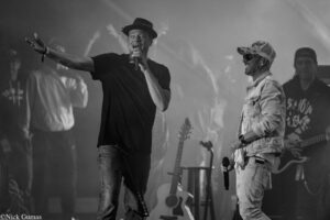 Two male singers perform on a smoky stage, one in a black T‑shirt singing into a handheld mic while the other in a denim outfit smiles with a mic in hand nearby.